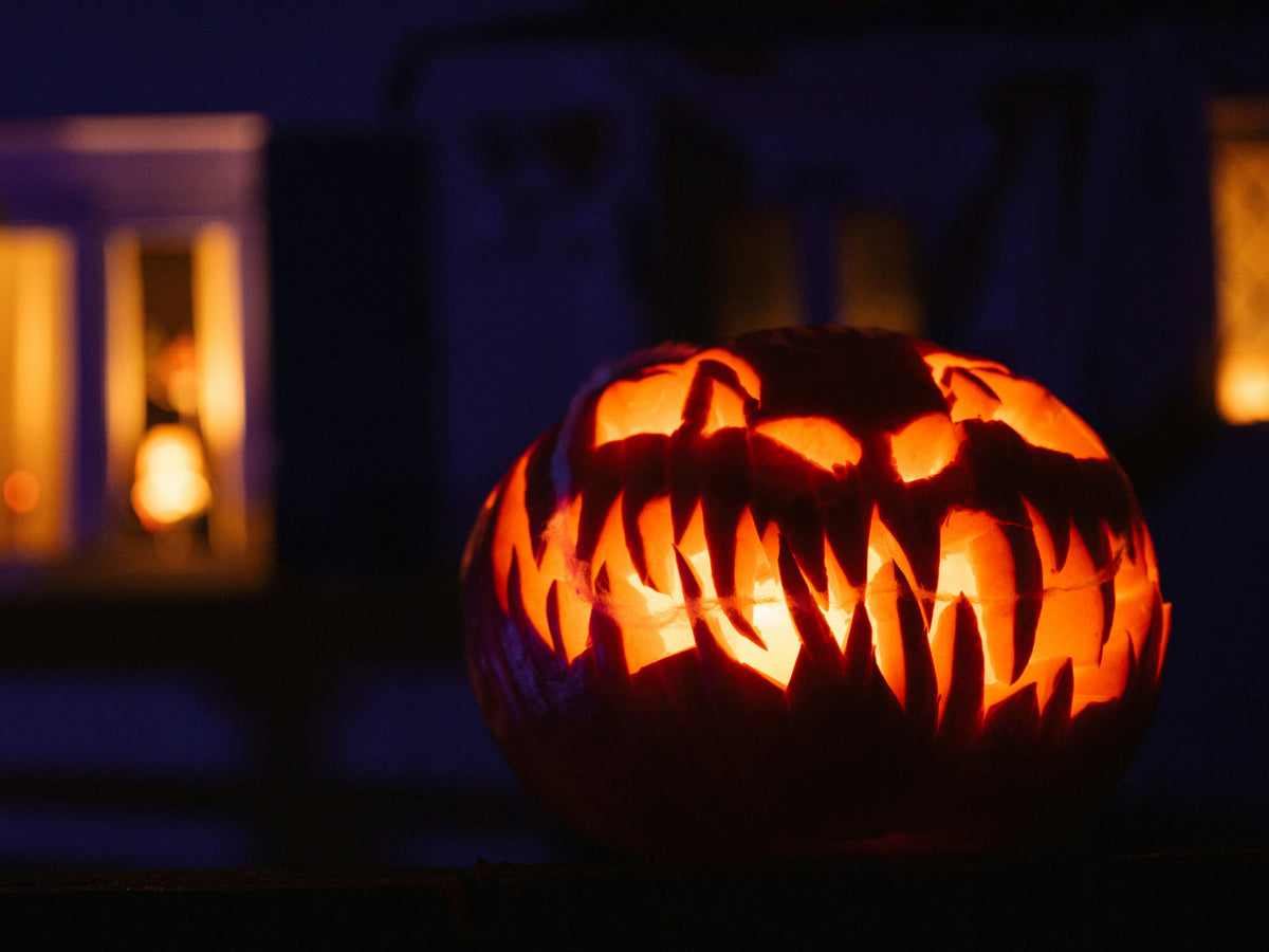A glowing carved pumpkin lantern at night with a warmly lit window behind it — the history of Halloween lanterns and the meaning of a light in the dark
