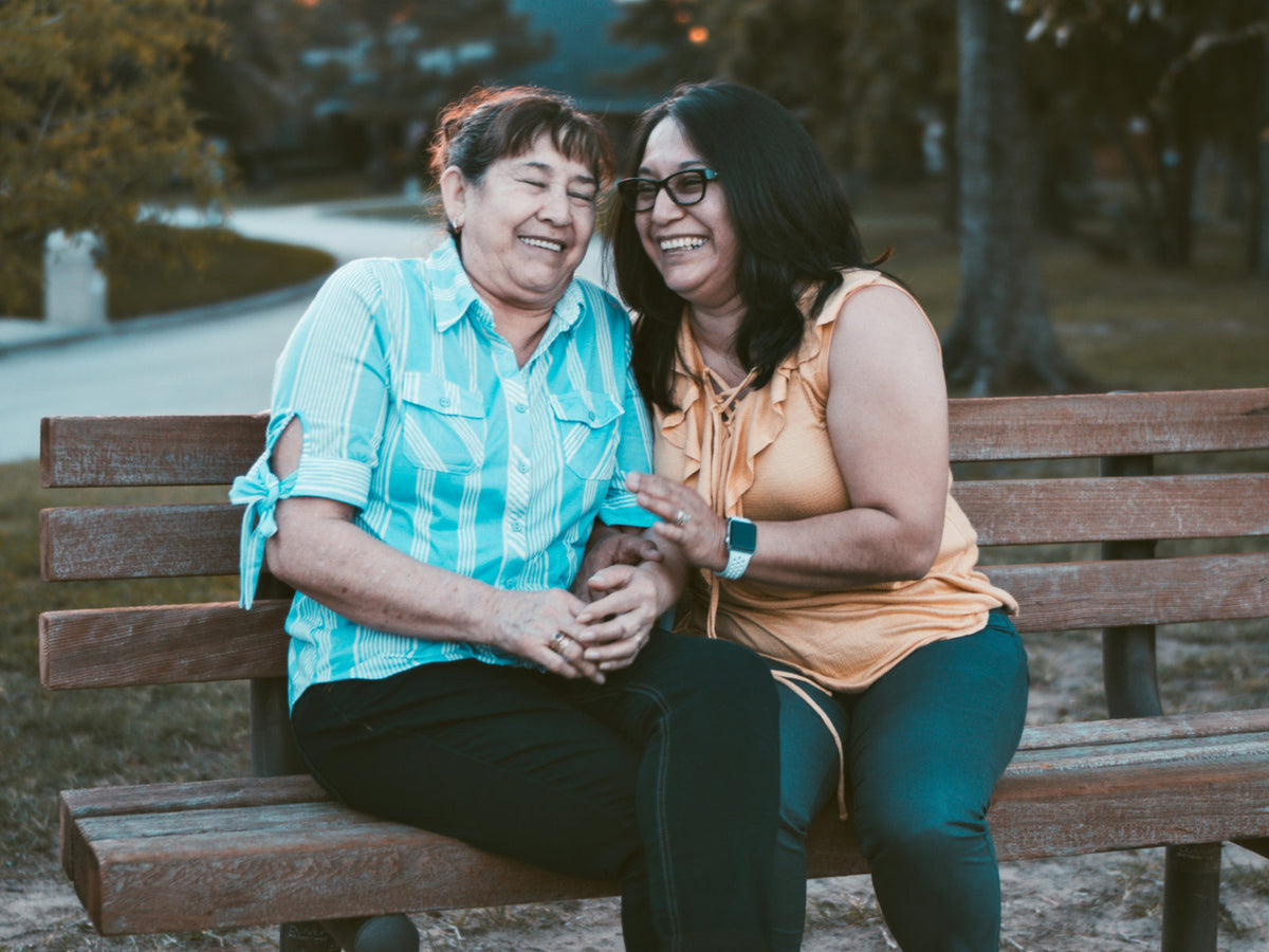 An older woman and a younger woman laughing together on a park bench at sunset — celebrating Mother-in-Law Day and the bond with the other mother in the family