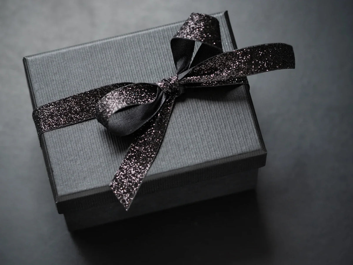 A black textured gift box tied with a sparkling black glitter ribbon on a dark background — representing the moment before opening, when elegant packaging already shows the care behind the gift