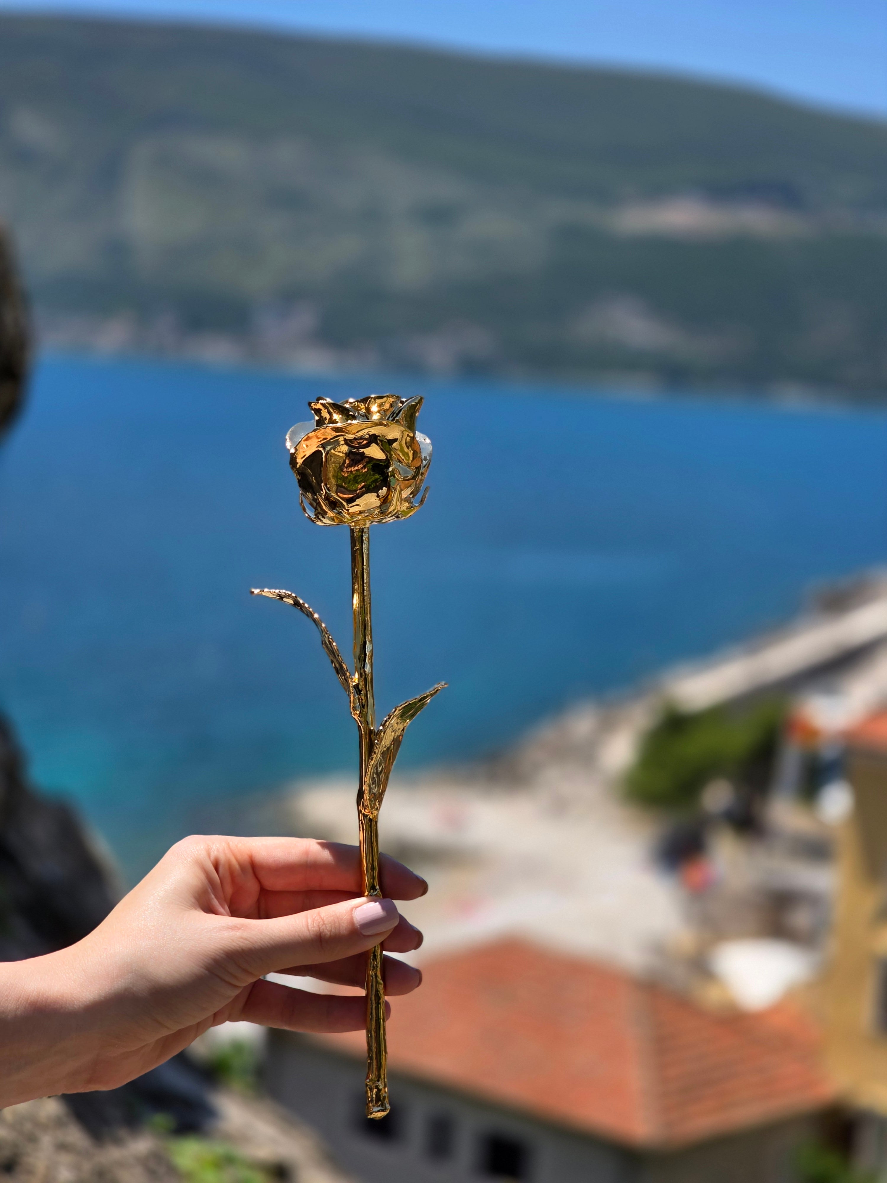 24K gold dipped rose held up against blue sea and coastal hillside view